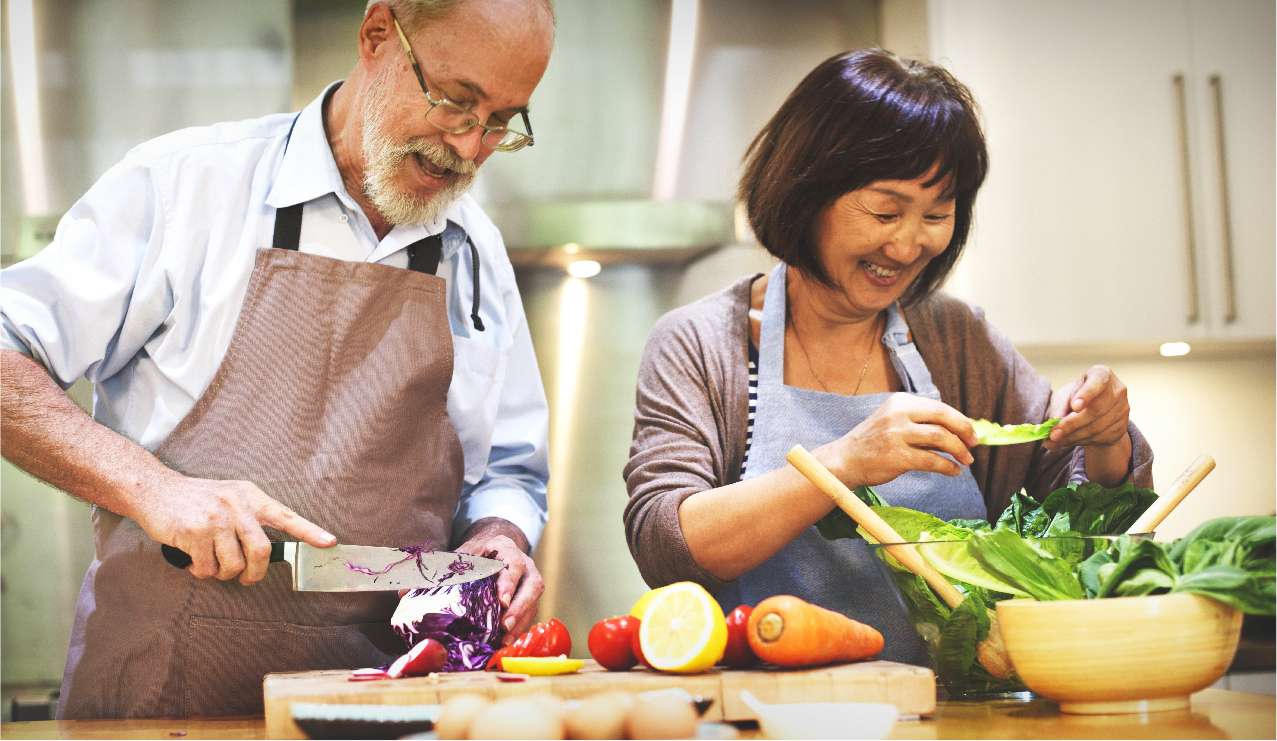 A senior man joyfully cuts a cabbage while a woman holds lettuce, both preparing a meal together in a kitchen.