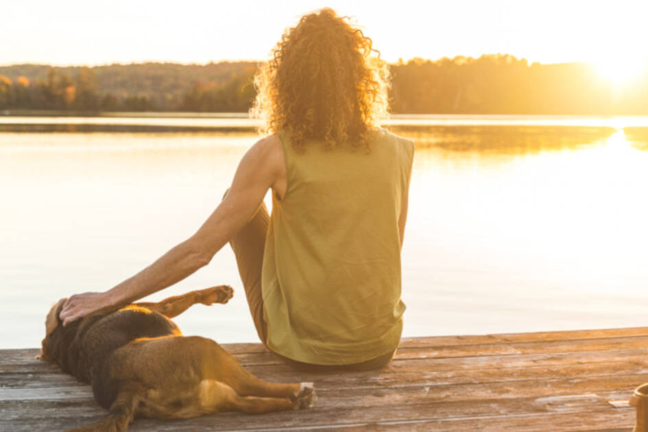 Anxiety and Depression - Lady with her dog sitting by a lake at sunset, the dog laying down next to her with her hand on its back, sharing a peaceful moment.
