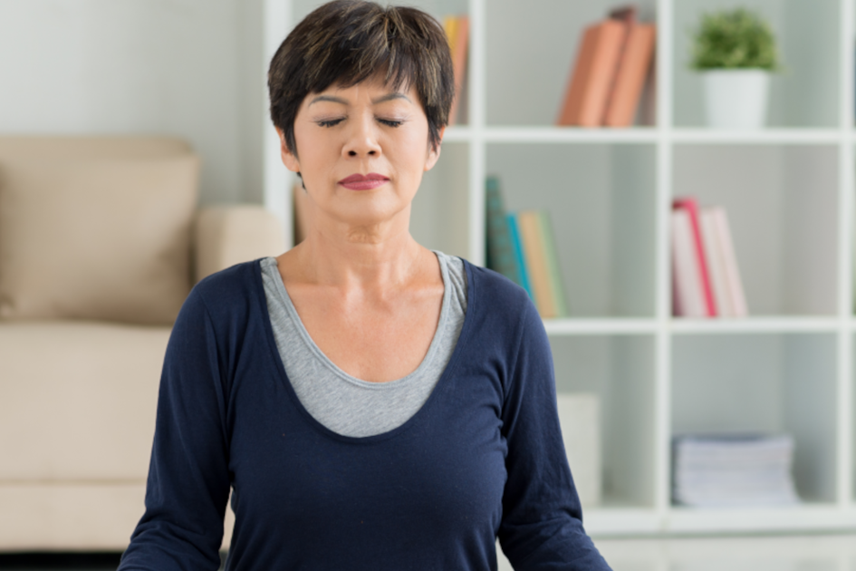 Middle-aged Asian woman sitting in meditation pose in her living room, practicing mindfulness exercises for blood pressure management.