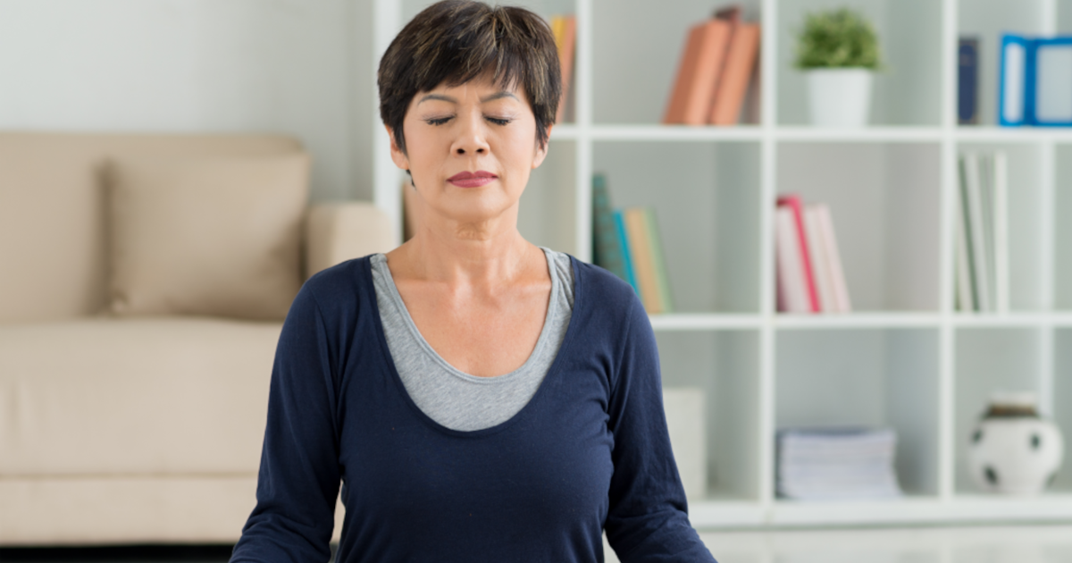 Stress Management - Middle-aged Asian woman sitting in meditation pose in her living room, practicing mindfulness exercises for blood pressure management.