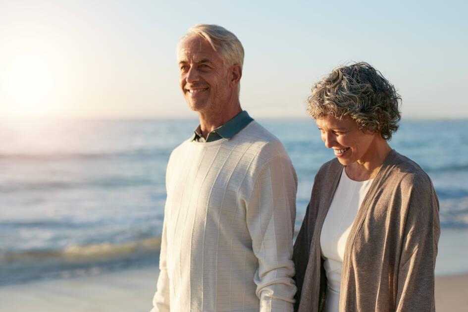 Happy senior couple, a lady and her husband in their 50s and 60s, walking hand-in-hand on the beach at sunrise, embodying vitality and heart health.