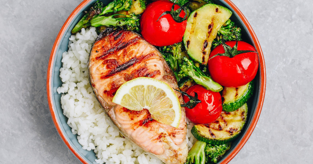 A nutritious meal showcasing the Mediterranean diet with white rice, grilled salmon, slices of avocado, tomato, and cucumbers on a plate.