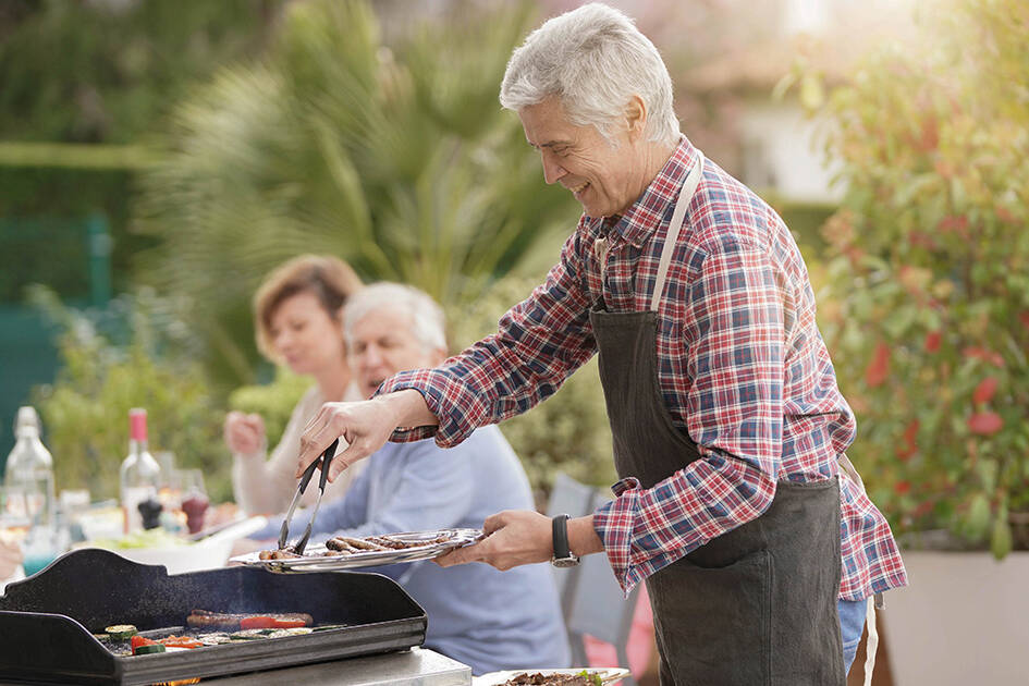 Community and Heart Health: Happy senior man grilling food on a barbecue with a woman sitting in the background, preparing to eat as a group outdoors during the day.